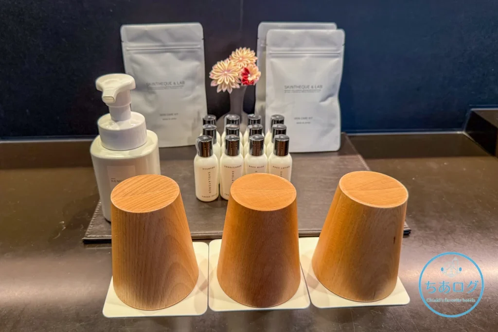 熱海パールスターホテルのパウダールームに用意されたSHOLAYERED（ショーレイヤード）のバスアメニティ、NEMOHAMOのハンドソープ、SKINTHEQUE & LABのスキンケアセットと温かみのある木製コップ。