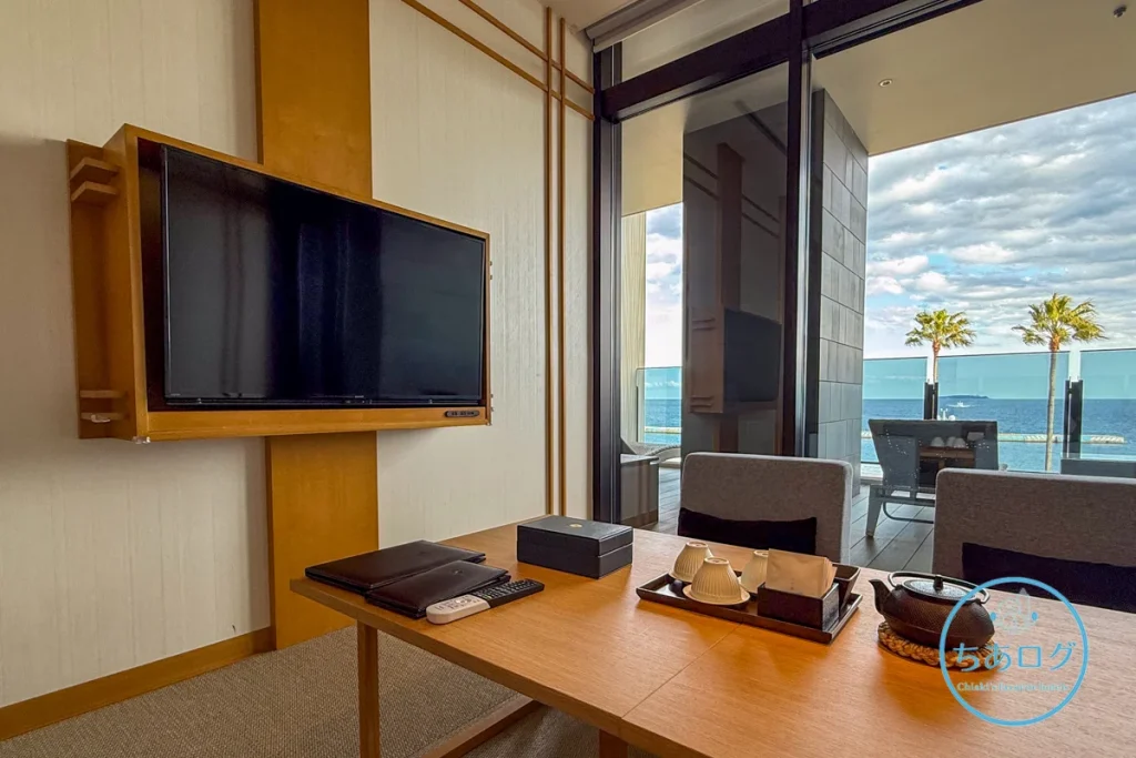 熱海パールスターホテル客室のリビングスペース。南部鉄器の急須と湯呑み、ぐり茶が置かれた低めの和テーブルと座椅子のセット。背景には熱海の海。