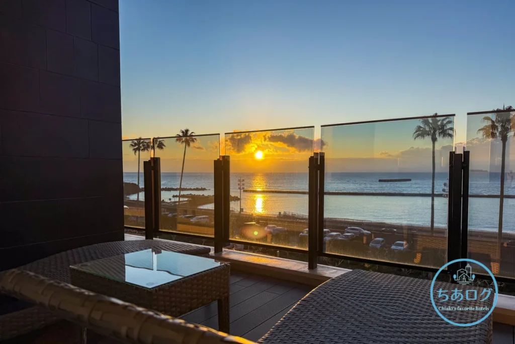 熱海パールスターホテル客室のテラス。ラタン調の寝椅子（サンラウンジャー）とテーブル、ガラス手すり越しに広がる熱海サンビーチの朝陽。